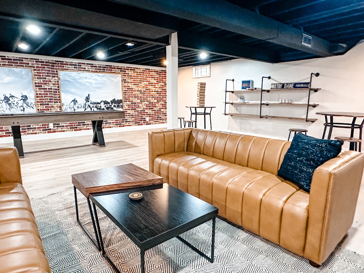 The basement hangout room features a spacious seating area with two brown leather sofas and a low coffee table. A shuffleboard game is visible against a textured brick wall, while additional high-top tables are arranged nearby for social gatherings.