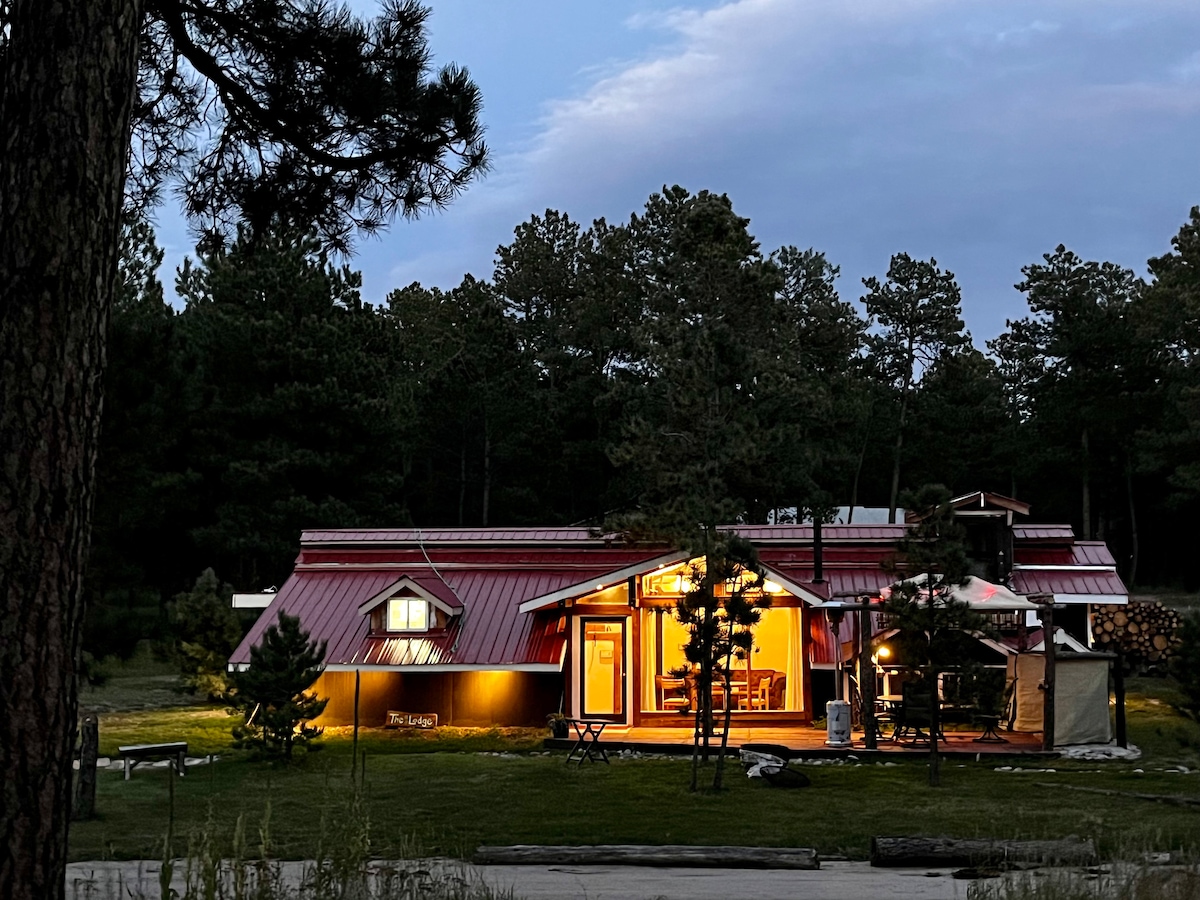 The lodge is illuminated in warm light against a twilight sky, featuring a distinctive red roof. Outdoor seating is visible, nestled among trees, with a patio umbrella providing shade. The inviting ambiance highlights the rustic charm of the surrounding natural landscape.