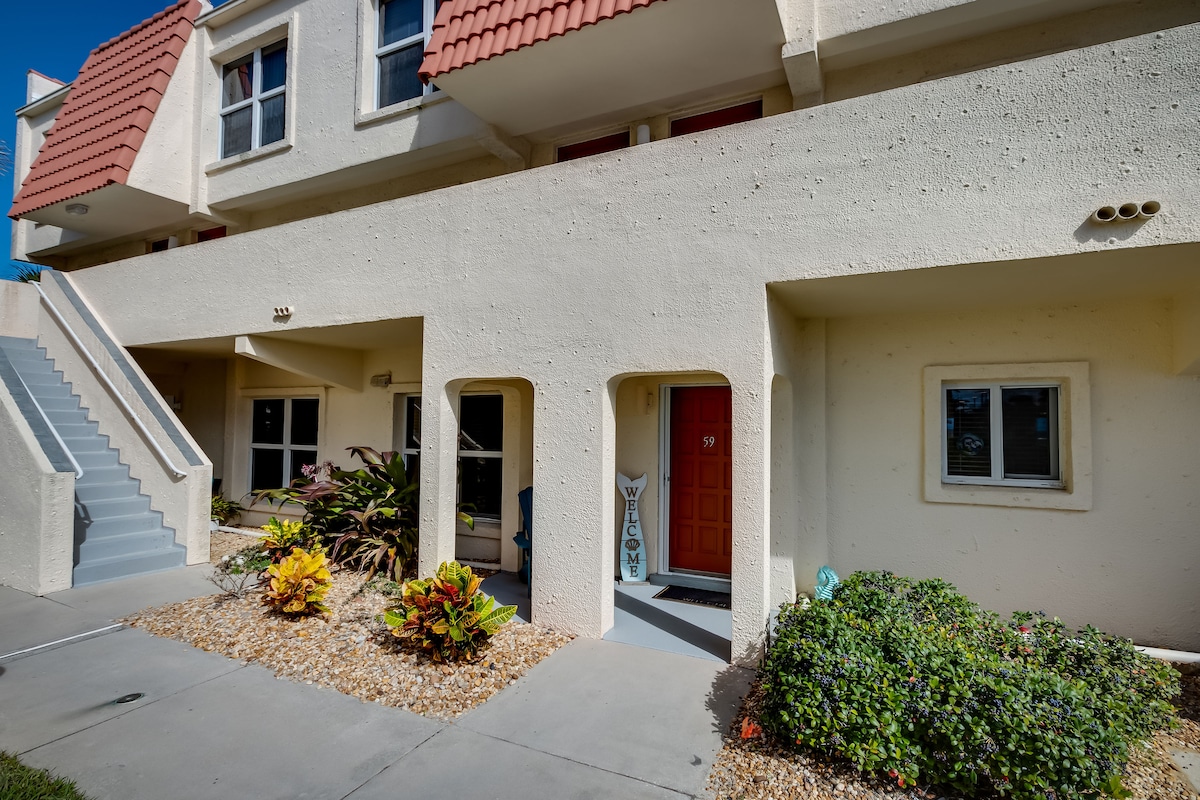 The exterior of the condo features a textured wall in pale tones with a red-tiled roof, surrounded by landscaped greenery. A set of stairs leads to the upper level. An inviting front door, numbered 59, is framed by vibrant plants, creating a welcoming entrance.