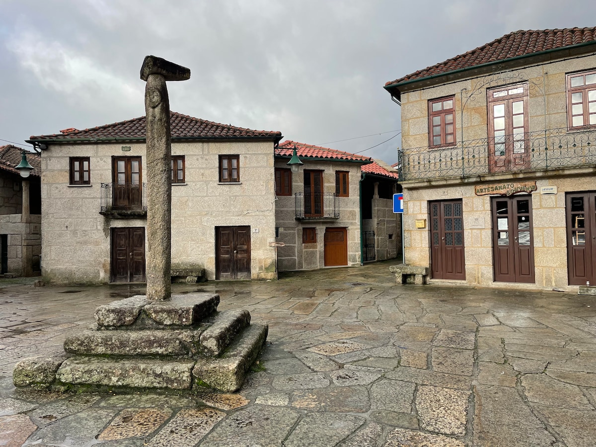 A stone pillar stands in the center of a quaint square, flanked by traditional granite buildings with wooden balconies. The surroundings feature cobbled pathways and a subtle overcast sky, creating a serene ambiance in this charming village setting.