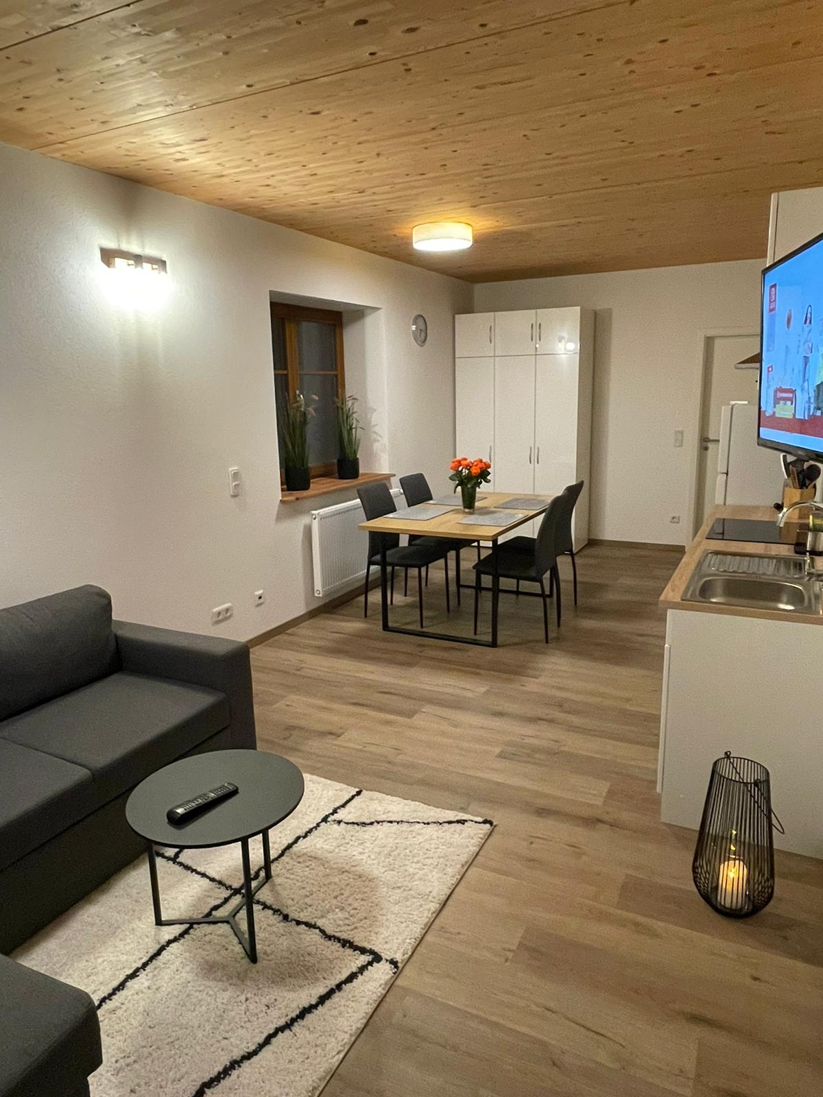 An inviting living area features a comfortable couch and a small coffee table, complemented by a patterned rug. A dining table with four chairs is visible, while a modern kitchen area is positioned in the background. Wall-mounted lighting enhances the warm atmosphere.