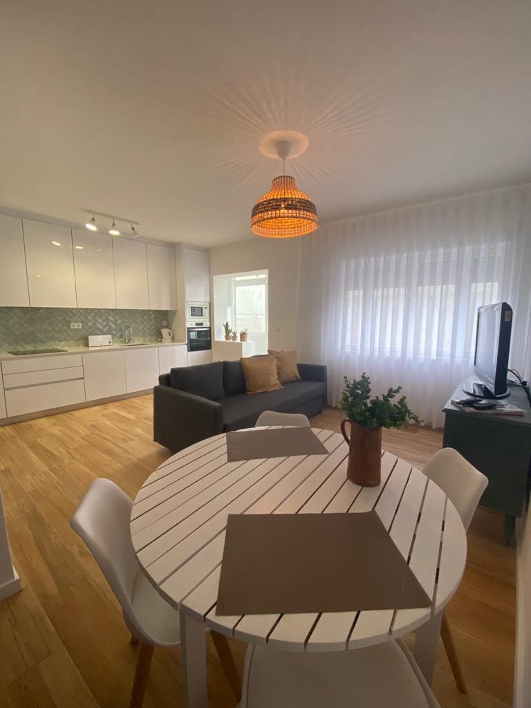 A modern living area features a gray sofa adorned with decorative cushions. A round white dining table with chairs is positioned nearby. Natural light fills the space through sheer window treatments, and a pendant light casts a gentle glow from above. A flat-screen television is visible on a side table.