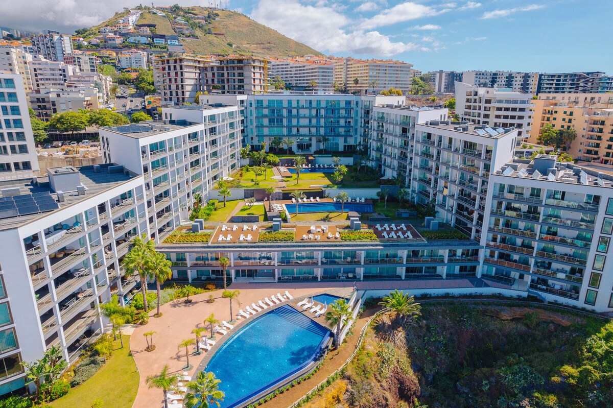 The aerial view captures the expansive layout of the Madeira Palace Residence, showcasing multiple buildings arranged around lush gardens and pools. A shimmering blue pool glistens in the center, framed by sun loungers. The surrounding landscape features hills and city buildings, illustrating the blend of natural and urban beauty.