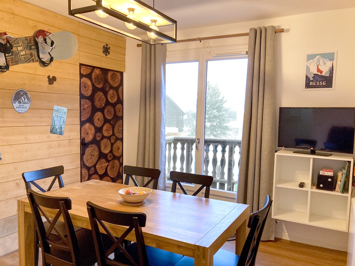A dining area is presented with a wooden table accompanied by black chairs. The window offers a view with sheer curtains, while a television and a white bookshelf are positioned nearby. Natural wood accents enhance the space, complementing decorative elements on the walls.