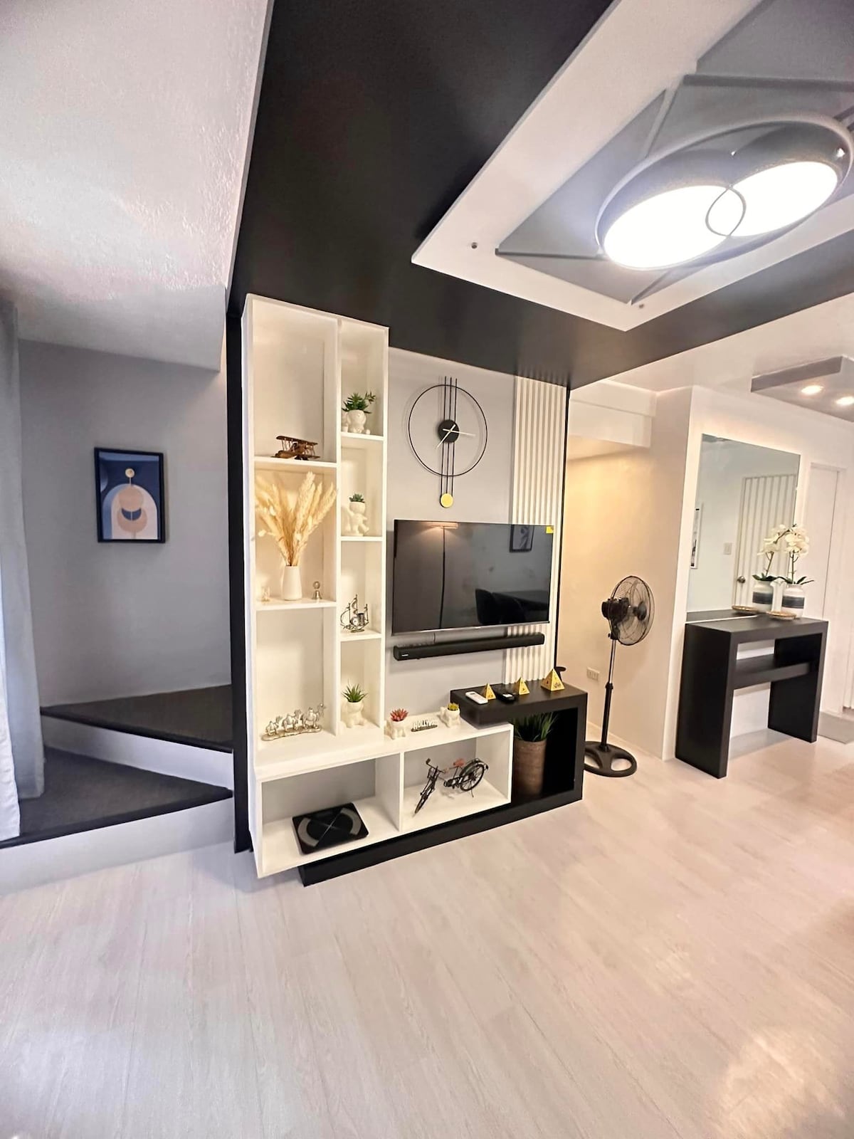 The living area features a wall-mounted TV and a stylish shelving unit, adorned with decorative items. A fan provides comfort, and a sleek table is positioned nearby. A staircase is visible in the background, leading to the private bedrooms on the upper floor.