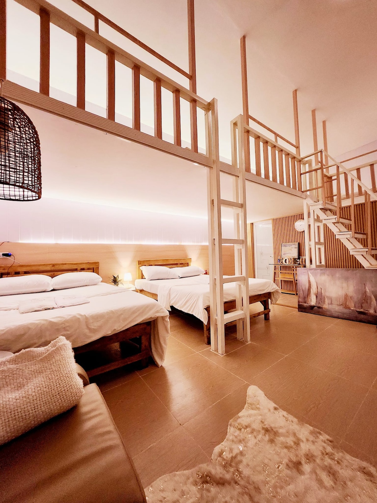 The spacious room features a wooden loft with twin beds and a queen-size bed below. Neutral-toned bedding creates a calm environment. A cozy seating area is included, and soft lighting highlights the warm wooden accents and minimal decor, enhancing the overall sense of comfort.