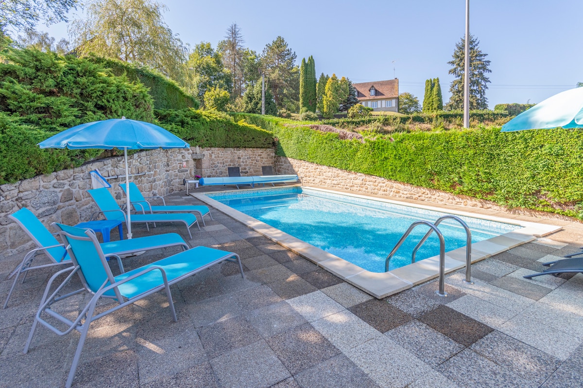 An outdoor swimming pool area is featured, surrounded by well-maintained greenery. Several blue lounge chairs are arranged neatly along the poolside, accompanied by light blue umbrellas. The crystal-clear water of the pool sparkles under bright sunshine, enhancing the tranquil setting.