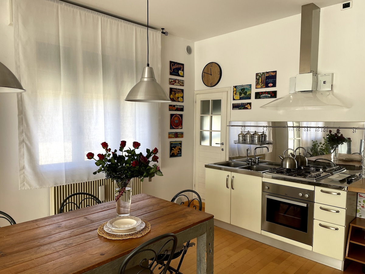 A light-filled kitchen features a wooden dining table set with a plate, surrounded by black metal chairs. A vase of red roses adds a touch of color. On the wall, framed artwork is displayed, and stainless steel appliances are visible in the cooking area.