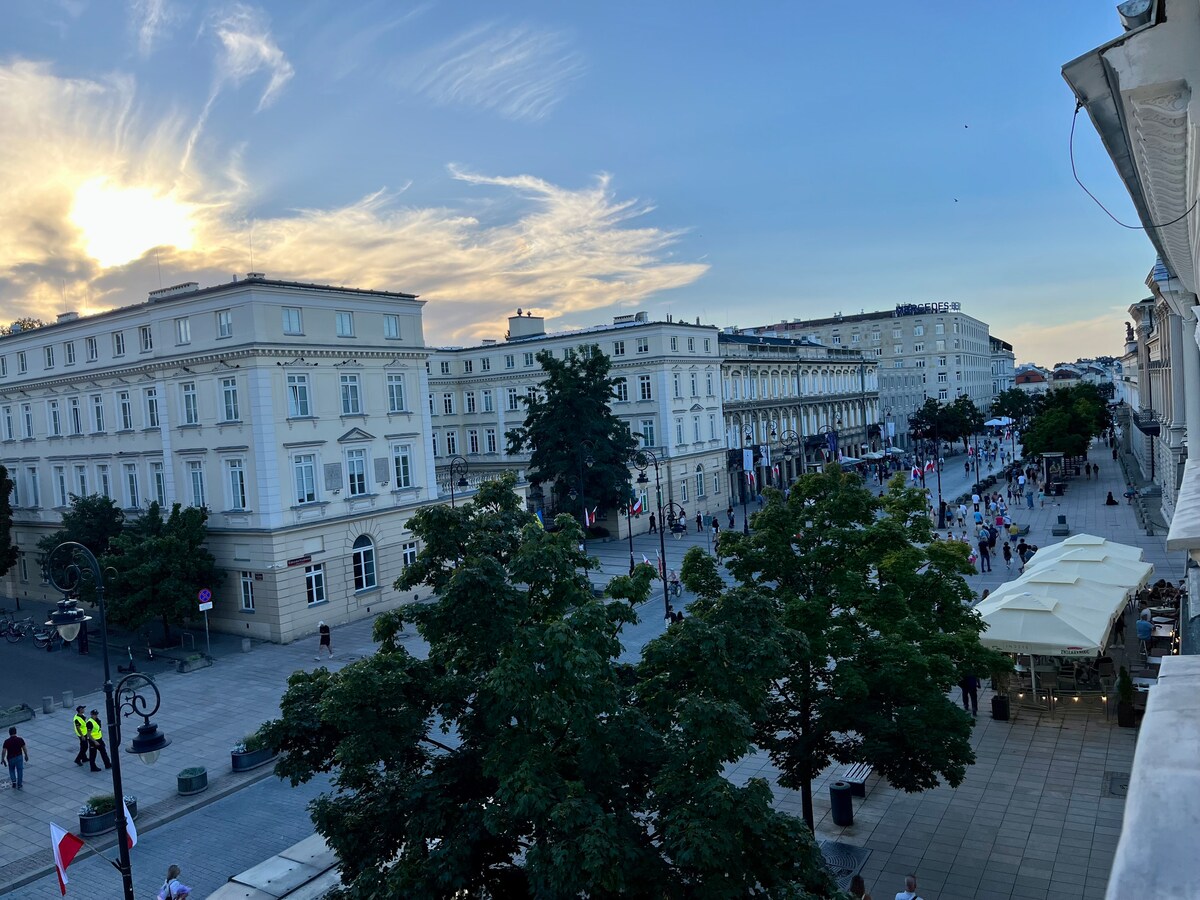 A view of a wide, tree-lined street populated with pedestrians, featuring elegant buildings on either side. The sky showcases soft hues as the sun sets, and outdoor seating with umbrellas is visible on the right, adding to the vibrant atmosphere of the area.