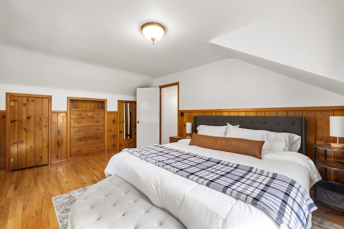 A spacious bedroom features warm wooden paneling and a cozy bed adorned with a soft blanket. A bedside table with a lamp complements the room, while a tufted bench sits at the foot of the bed. Ample natural light highlights the serene atmosphere.