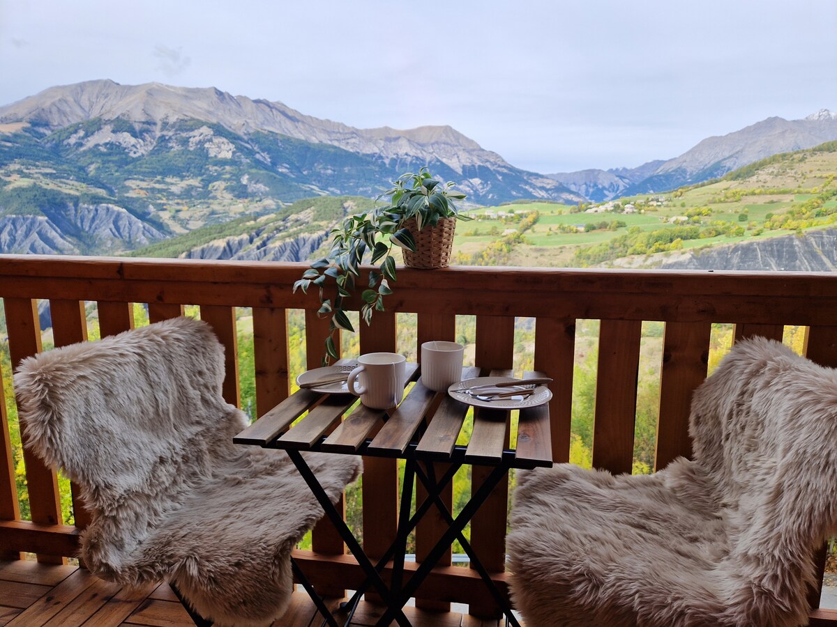 A cozy balcony setting is presented with a small wooden table and two fur-covered chairs. A potted plant adds a touch of nature, while the backdrop features a sweeping panoramic view of the valley and mountains, enhancing the serene outdoor experience.
