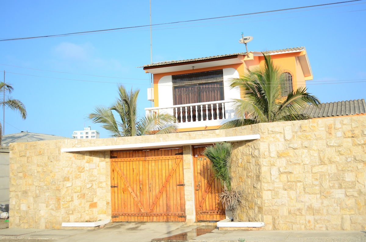 A two-story house showcases a blend of traditional architecture and coastal charm, framed by palm trees. The front features a stone wall with wooden double gates leading to the entrance. A balcony is visible on the upper level, complementing the exterior's warm tones.