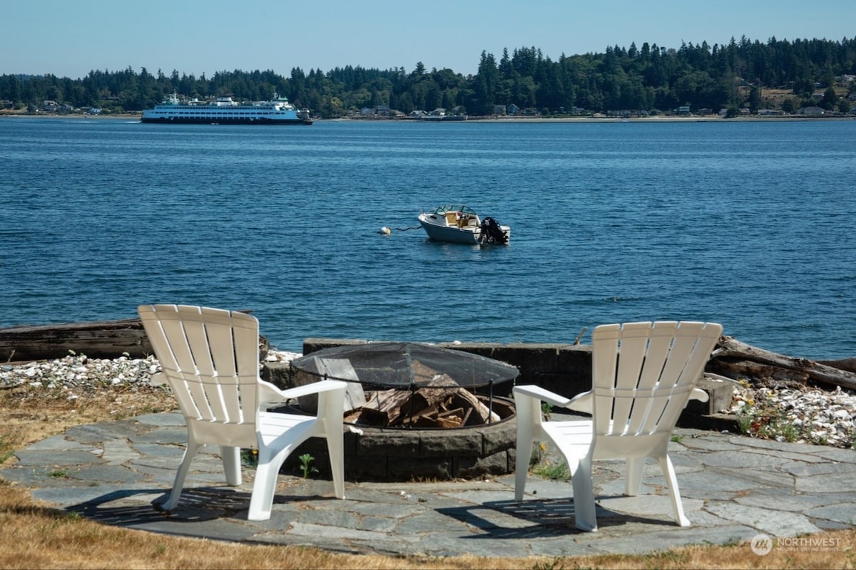 A tranquil fire pit area is framed by two adirondack chairs, set against a backdrop of calm water. In the distance, a ferry traverses the bay, while a small boat floats nearby. The surrounding shoreline is accented with pebbles and driftwood.