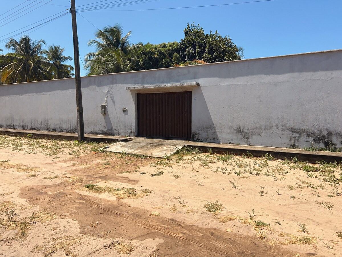 A wall surrounds the property, providing privacy, with a central wooden gate that leads inside. The ground features sandy terrain, and palm trees are visible in the background under a clear blue sky.