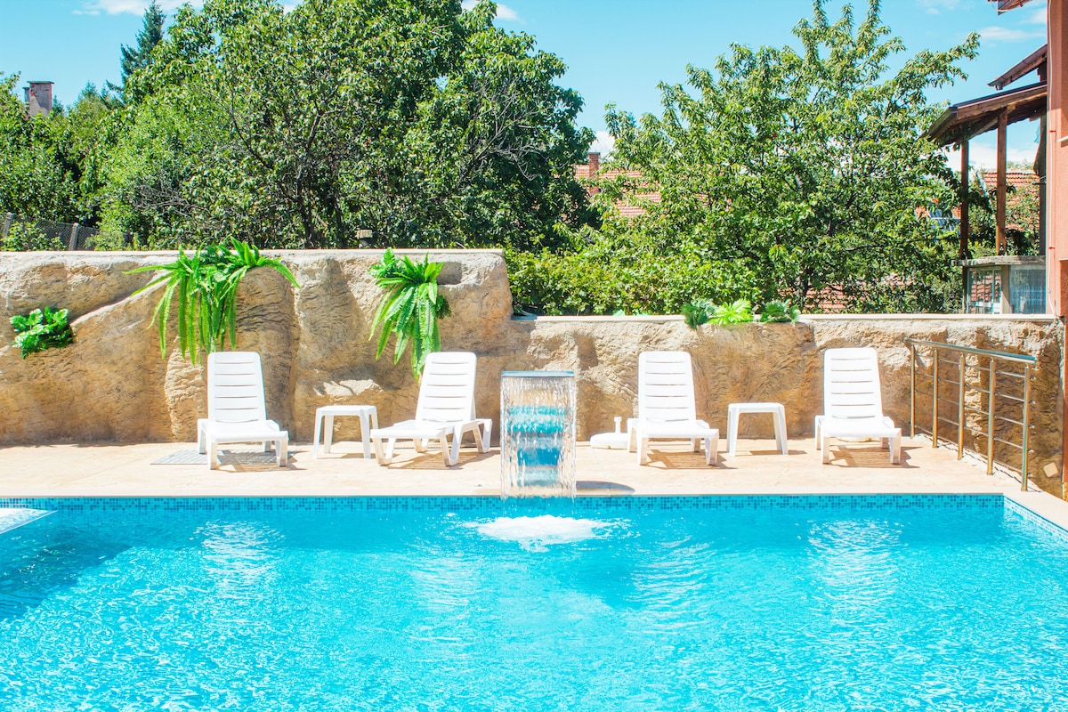 An inviting outdoor swimming pool features clear blue water and is surrounded by a stone wall adorned with greenery. Four white lounge chairs are positioned nearby, providing a relaxing spot for sunbathing, while a waterfall feature adds a serene touch.