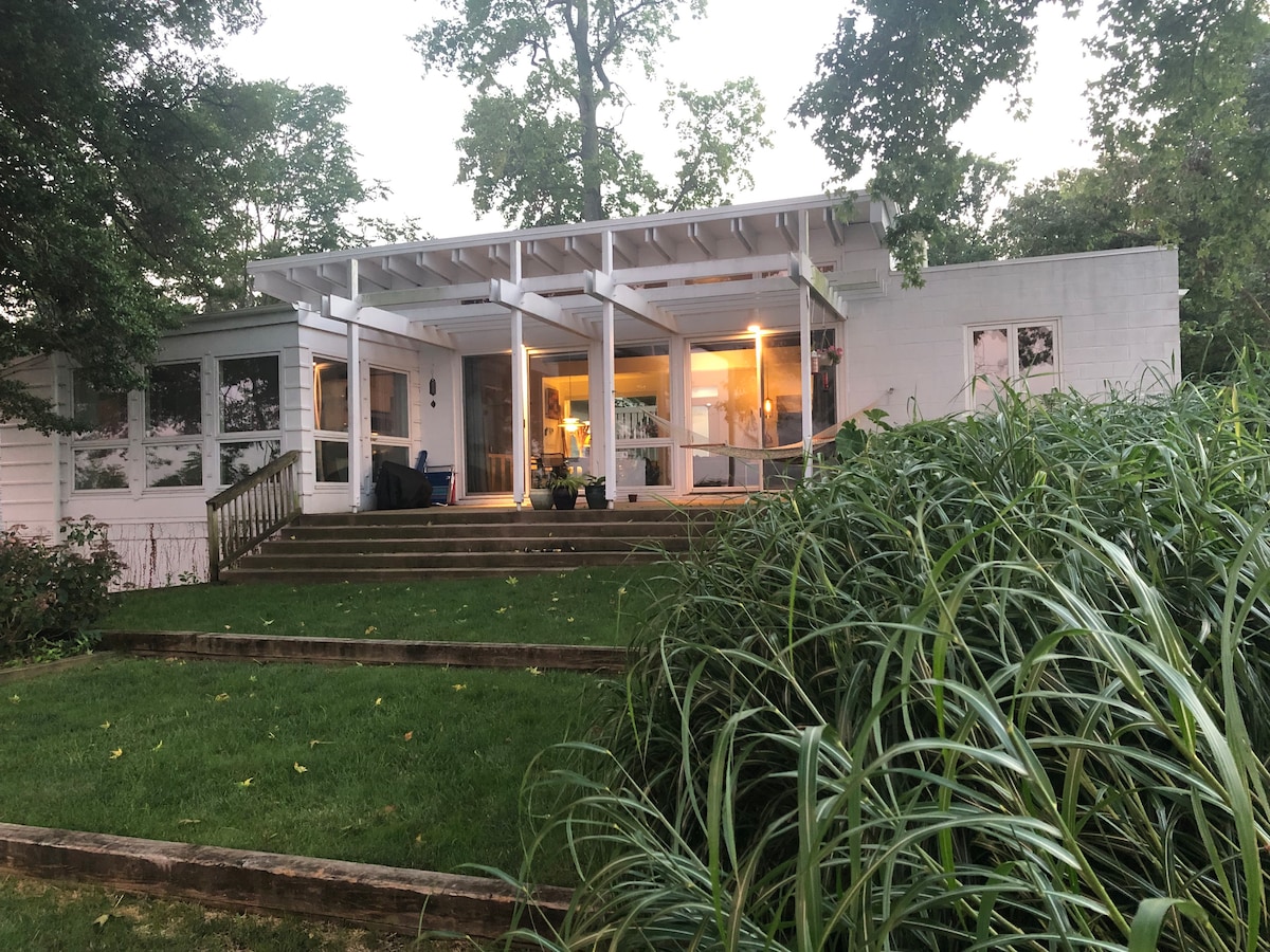 A modern house is set amidst lush greenery, featuring a spacious deck with wide stairs leading to the entrance. Large windows and a covered porch allow natural light to illuminate the inviting interior, creating a serene connection with the surrounding nature.