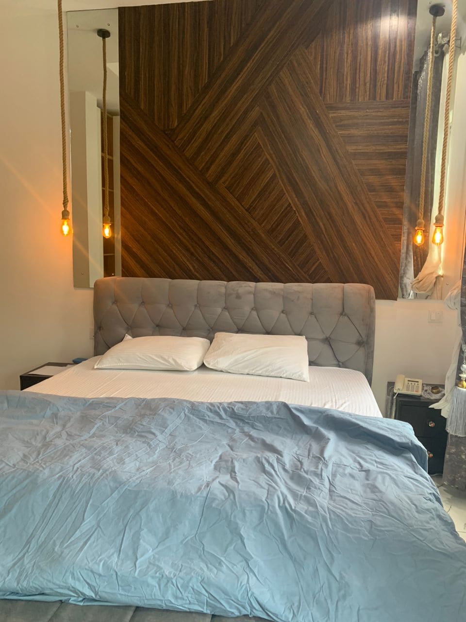 A spacious sleeping area is featured with a plush, upholstered bed and soft white linens. A textured wooden wall adds depth, while modern pendant lights hang above, providing gentle illumination.