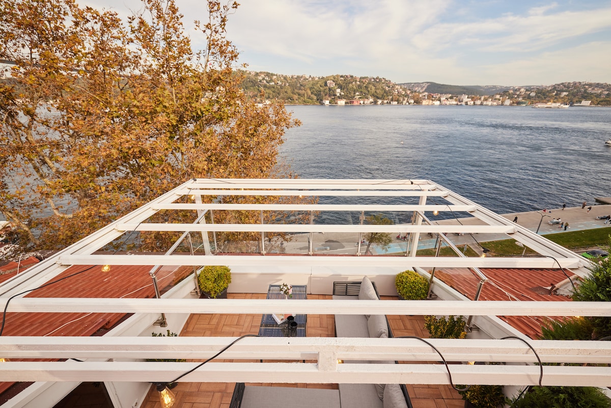 An outdoor terrace is highlighted, featuring seating areas and plants surrounded by a wooden deck. The stunning view of the water and hillside is visible, providing a serene backdrop with clear skies and subtle colors.