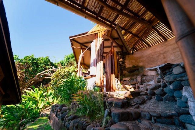 The exterior of a charming cabin is framed by vibrant greenery, featuring a thatched roof and natural stone steps leading to the entrance. Sunlight illuminates the pathway, creating a welcoming atmosphere surrounded by a blend of nature and rustic architecture.