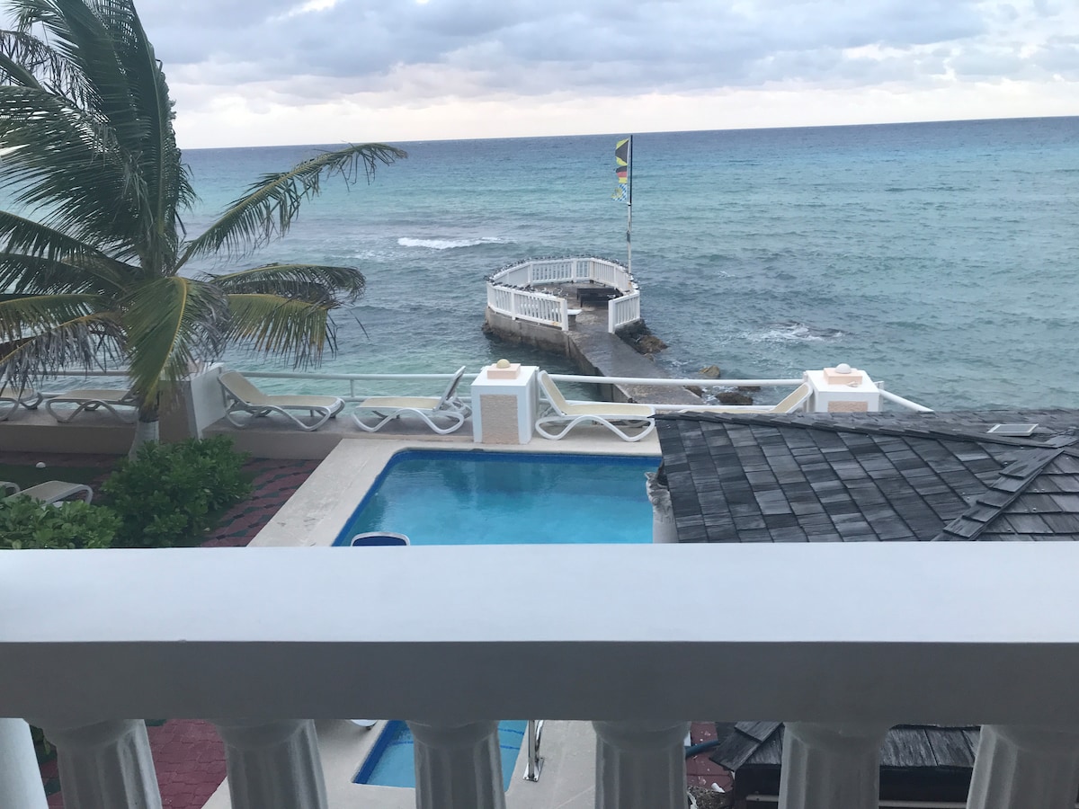 A scenic view of the ocean is presented from a balcony. A pathway extending into the water is visible, bordered by palm trees and lounge chairs. A swimming pool is situated nearby, reflecting the hues of the sea and sky.