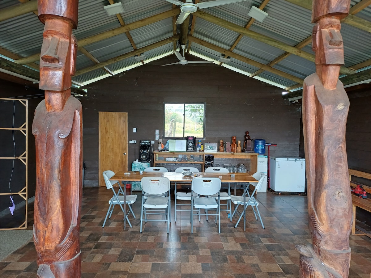 An open communal area is framed by two carved wooden figures. A large wooden table is surrounded by several white chairs. Various artifacts are displayed along the walls, complemented by a window that allows natural light to illuminate the space.