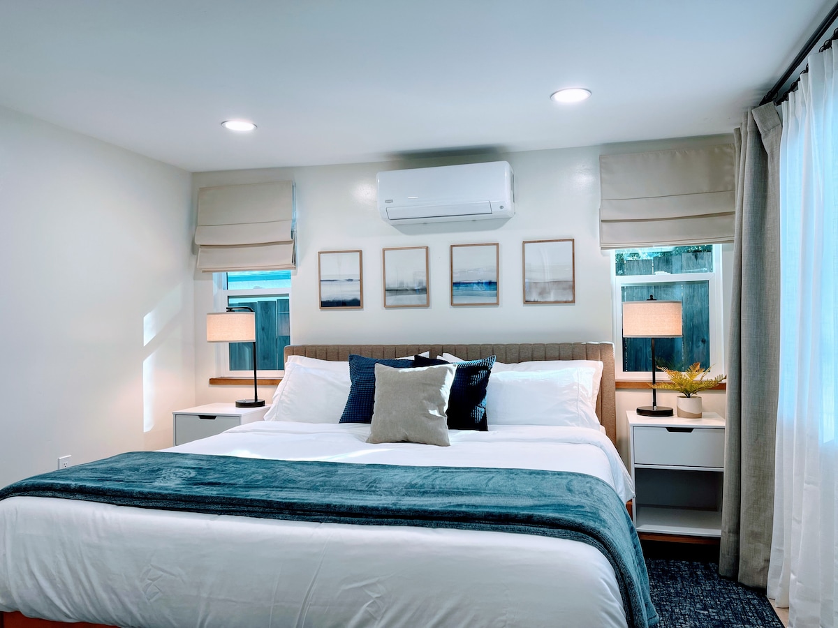 A well-appointed master bedroom features a spacious bed layered with soft linens and decorative pillows. Two bedside tables with lamps flank the bed, while cohesive artwork adorns the wall above. Natural light filters through the adjacent window, enhancing the tranquil ambiance.
