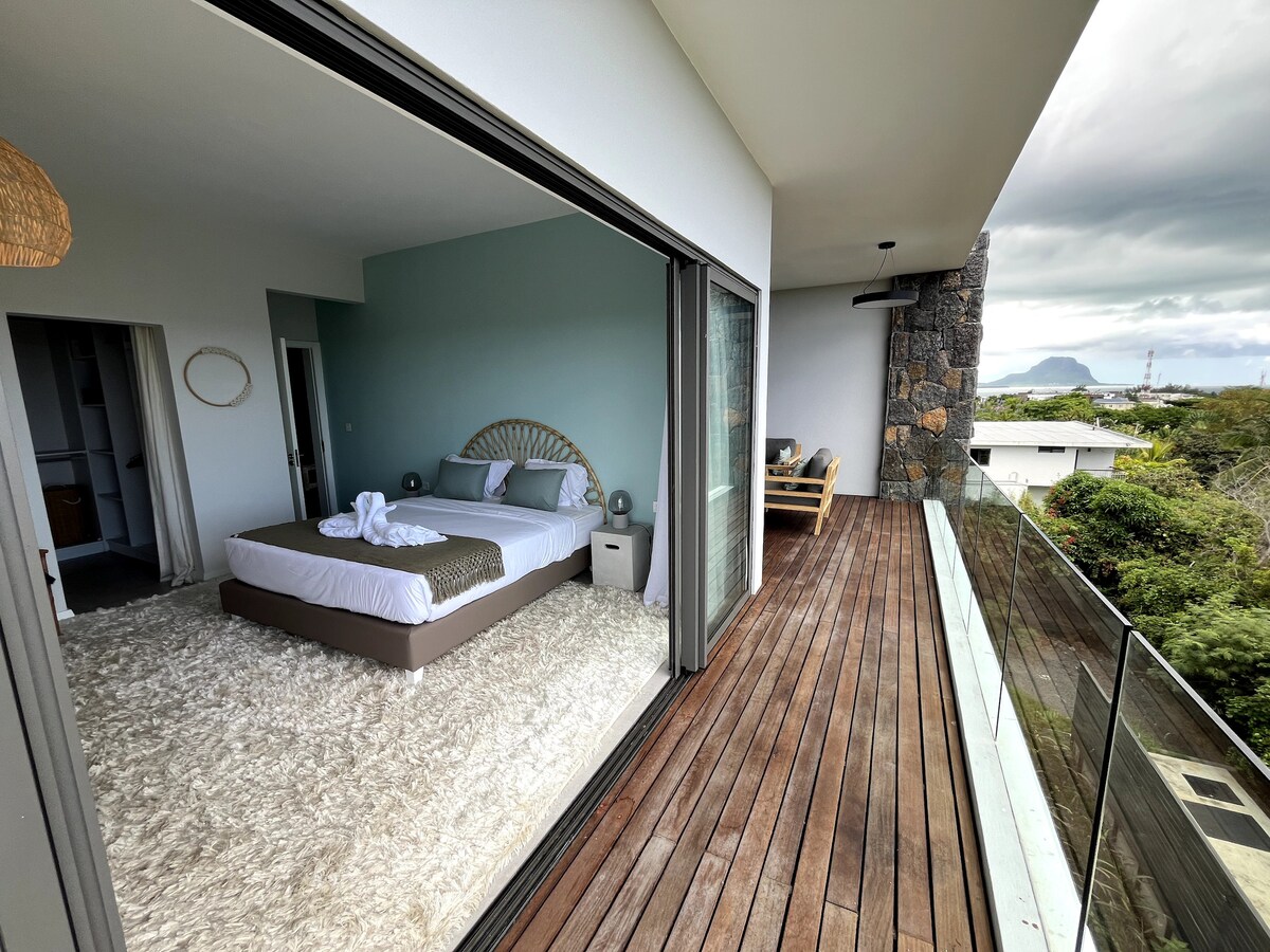A spacious bedroom features a comfortable bed, complemented by soft linens and a decorative headboard. Large glass doors open to a wooden terrace, offering scenic views of the surrounding greenery and distant mountains. A cozy seating area is visible, inviting relaxation.