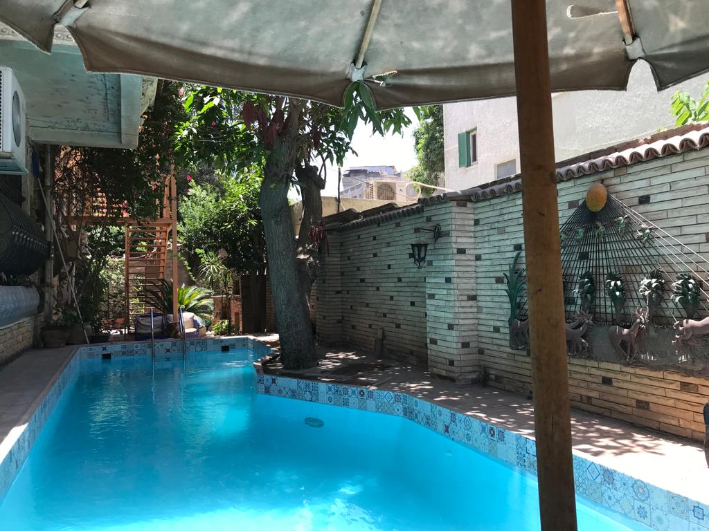 A well-maintained swimming pool is surrounded by lush greenery, with a shaded seating area provided by a large umbrella. The pool is accented by decorative tiles, and nearby foliage adds to the serene atmosphere of the outdoor space.