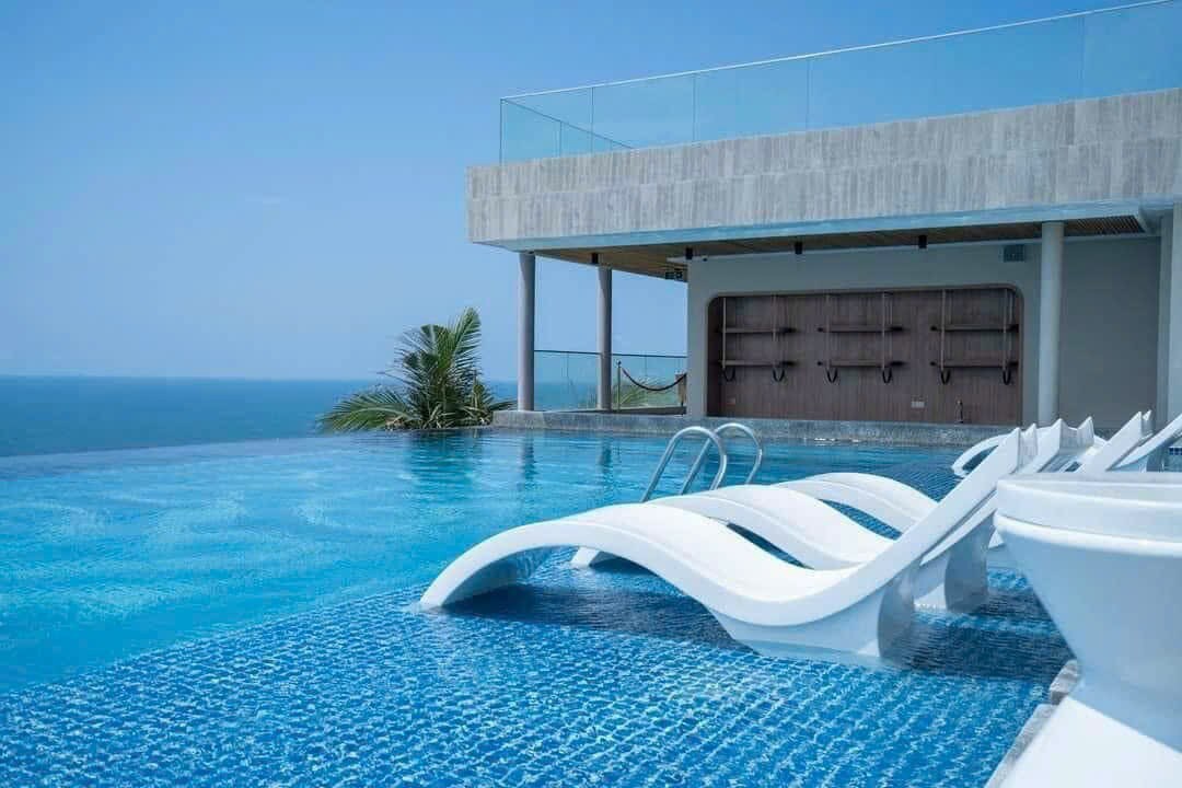 An infinity pool is situated overlooking the ocean, featuring sleek lounge chairs for relaxation. The area is bathed in natural light, with a clear blue sky above, and the distant horizon provides a serene backdrop.