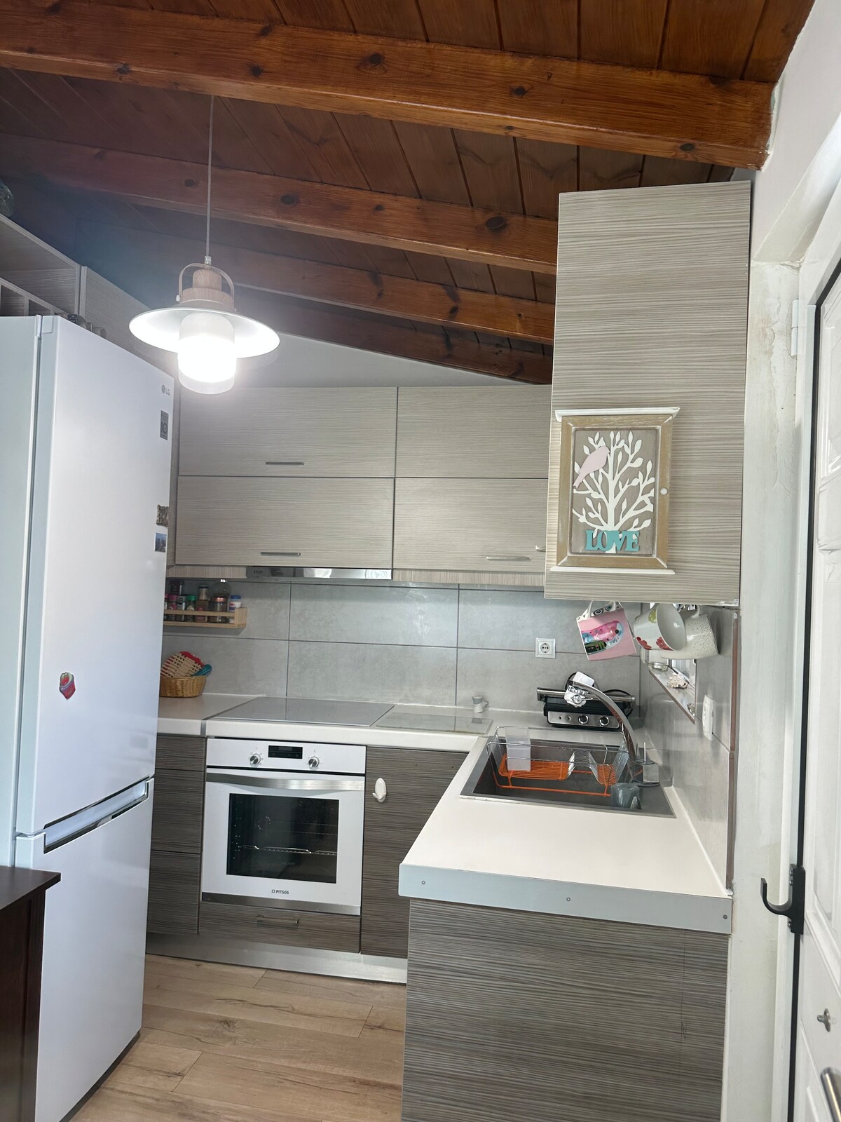 The kitchen features modern cabinetry with light wood accents and a clean countertop. A refrigerator and an oven are present, alongside a sink and cooking appliances. A pendant light hangs from the ceiling, illuminating the space, which is complemented by tiled walls and a wooden ceiling.