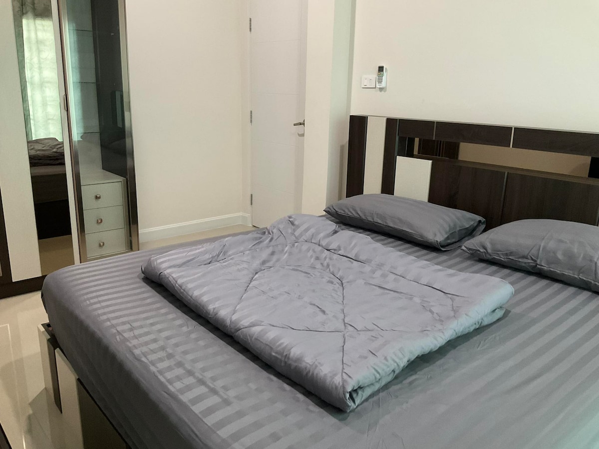 A spacious bed is presented with a light gray quilt neatly folded at the foot. The room is softly illuminated by natural light filtering through sheer curtains. A mirrored wardrobe is visible in the background, along with a hint of a door leading to another space.