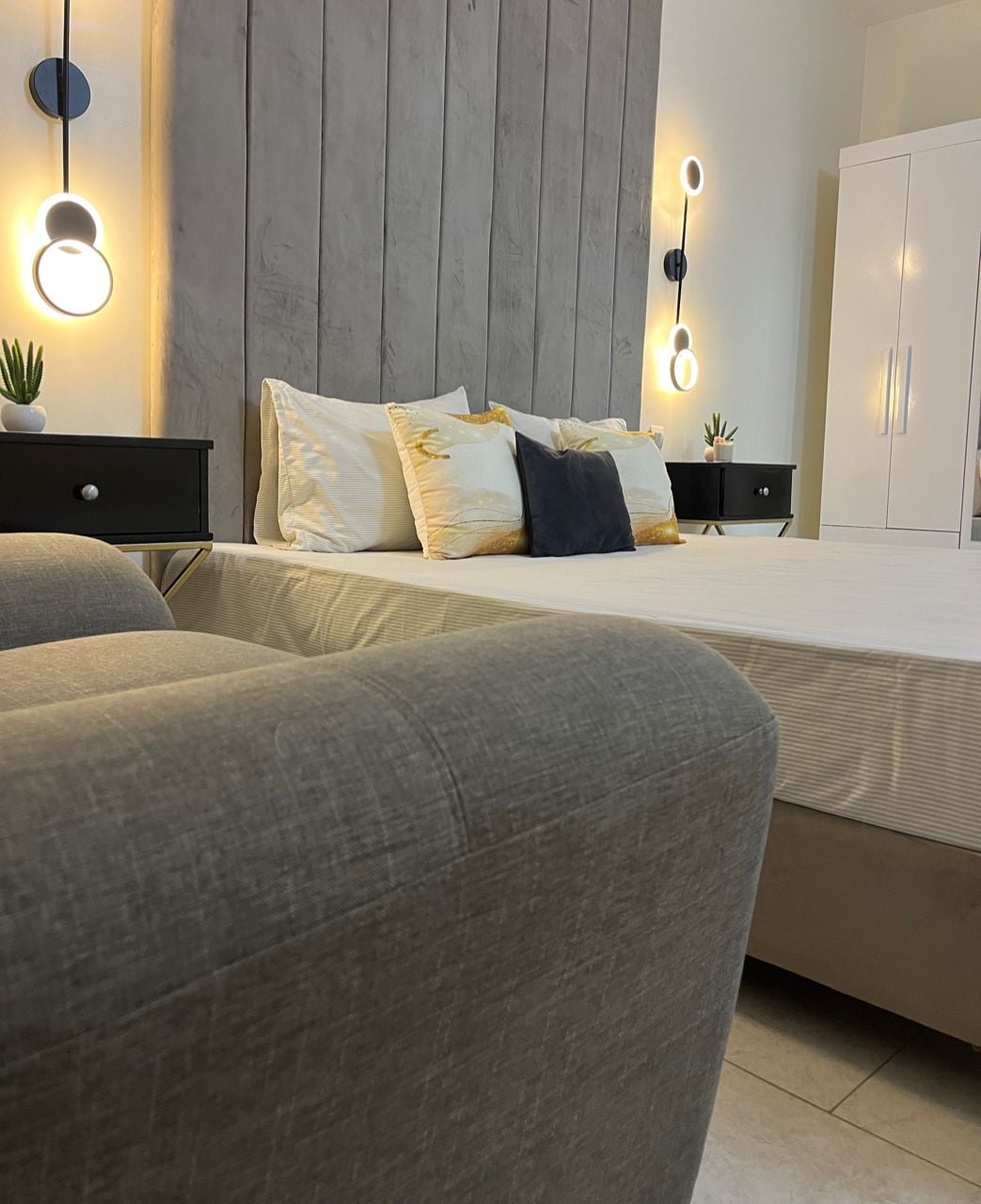 A spacious bed is positioned against a textured wall, flanked by two bedside tables featuring modern lighting. A cozy seating area is visible in the foreground, with neutral-toned upholstery. Decorative pillows add a subtle touch of color to the bedding.