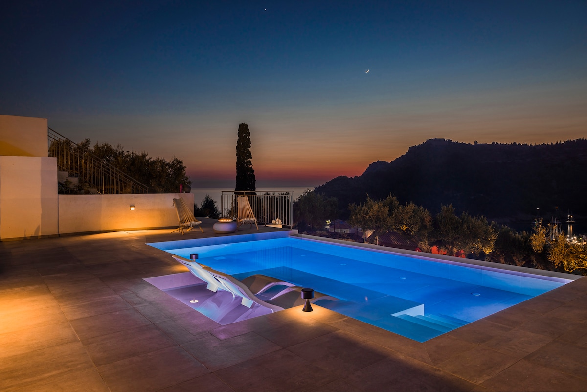 A modern pool area is illuminated in soft blue light, featuring gentle lighting around the edges. The pool offers a sleek, minimalistic design, and the surrounding landscape includes distant hills and a sunset sky reflecting hues of orange and purple.