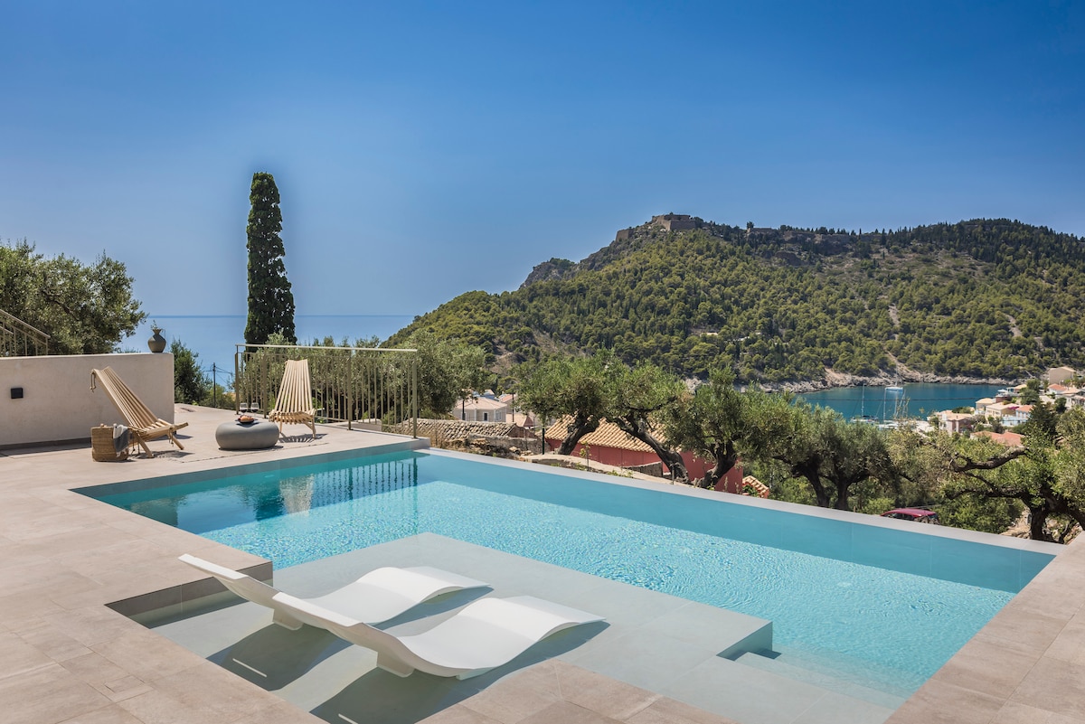 A serene outdoor space features a sleek infinity pool overlooking a scenic landscape of hills and the sea. Sun loungers are positioned nearby for relaxation under clear blue skies. Olive trees frame the area, enhancing the natural ambiance.