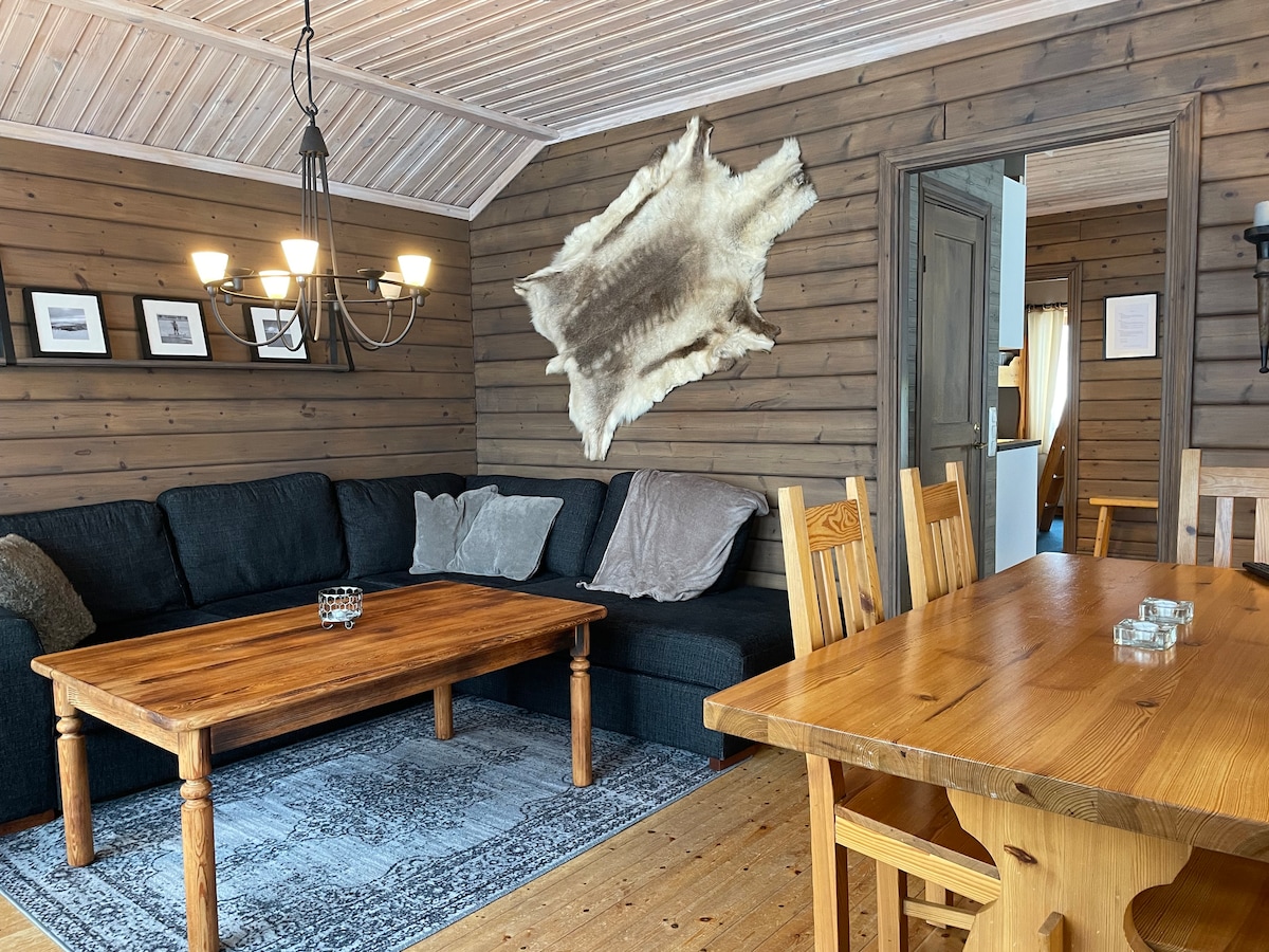 A cozy interior is featured, with a dark sectional sofa positioned around a wooden coffee table. A rustic dining table with wooden chairs is visible, alongside a wall displaying black-and-white photographs. Natural light enters through an open doorway, complementing the wooden paneling.