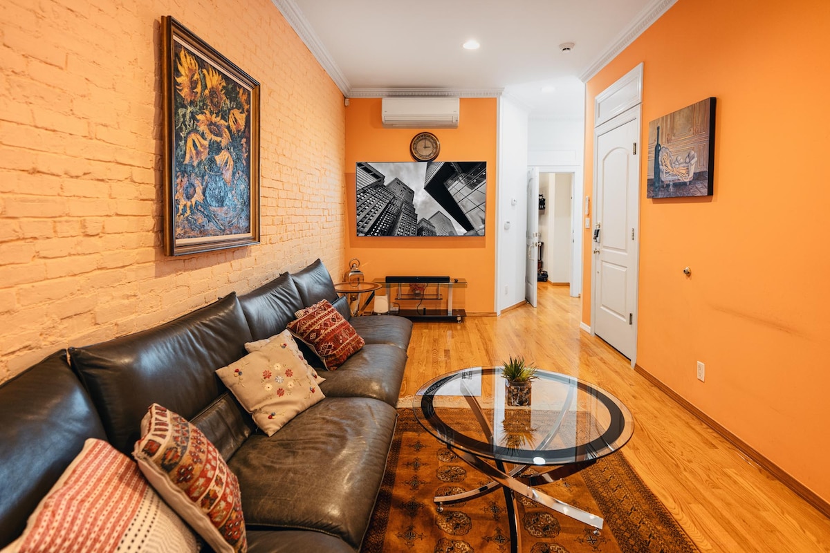 A spacious living room features an orange brick wall complemented by a large black leather sofa adorned with decorative pillows. A glass coffee table sits on a patterned rug, and modern art pieces decorate the walls. Natural light illuminates the area, enhancing its welcoming ambiance.