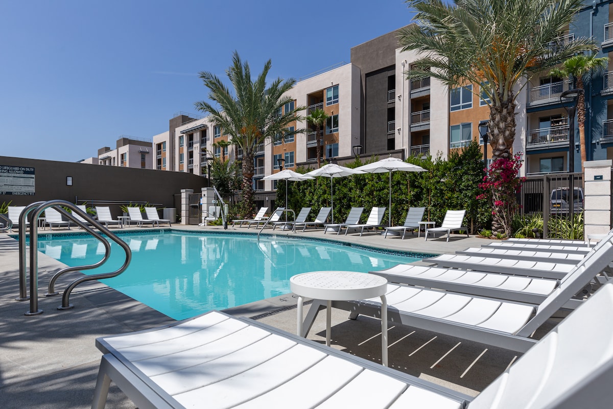 The outdoor pool area is surrounded by lounge chairs and umbrellas, offering a relaxing space for guests. Lush palm trees line the periphery, and the modern apartment complex is visible in the background, showcasing a blend of vibrant landscaping and contemporary architecture.