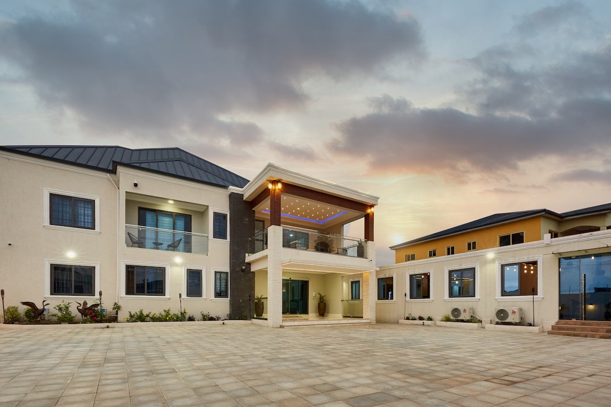 An expansive outdoor area features a stone-paved courtyard surrounded by the inn's modern architecture. The two-story building displays large windows and a covered terrace adorned with soft lighting, creating a serene environment at dusk. Lush greenery can be seen on the property.