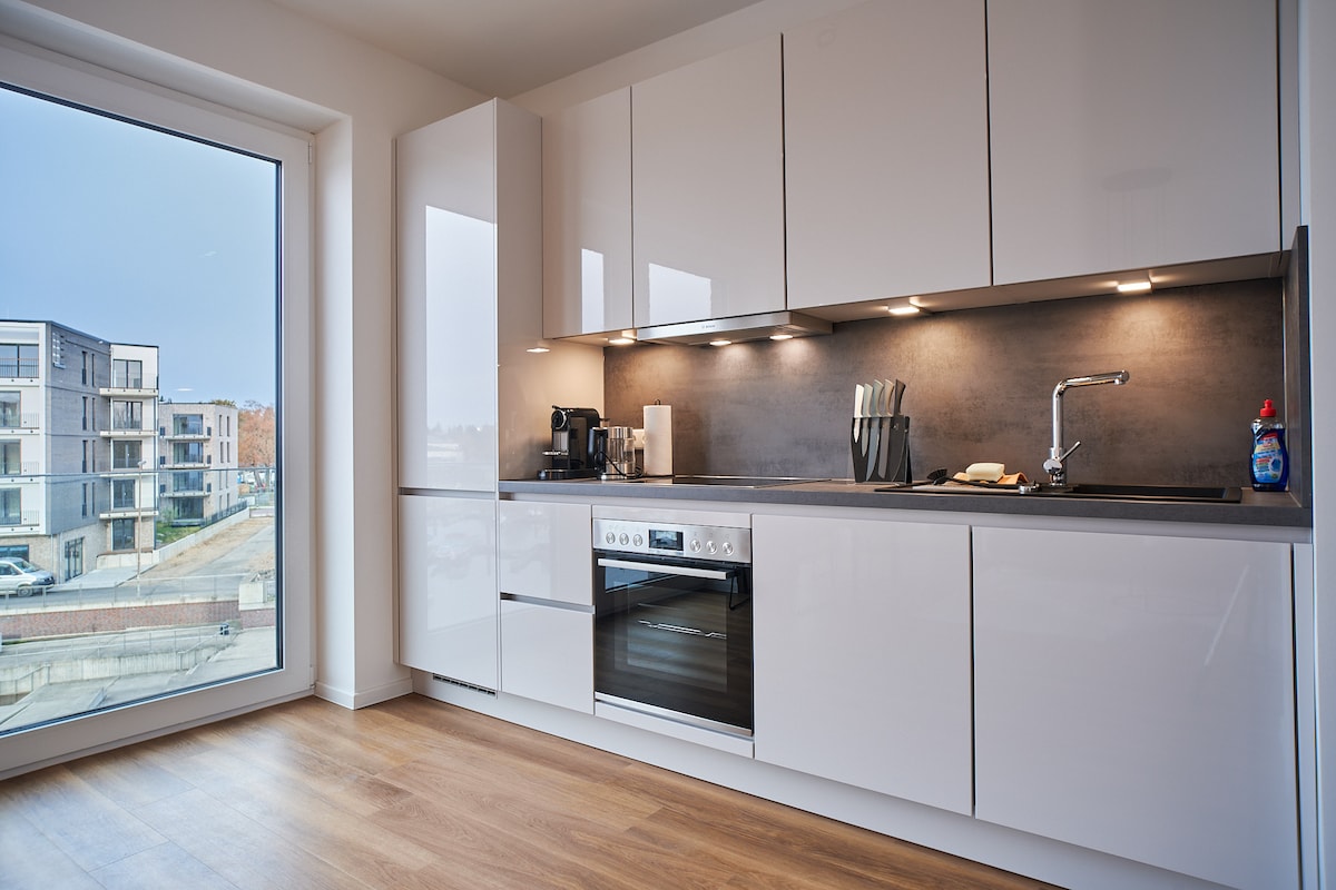 A modern kitchen is presented with sleek white cabinetry and a dark countertop. An oven and a coffee maker are visible, along with various utensils. A large window allows natural light, showcasing a view of nearby buildings.
