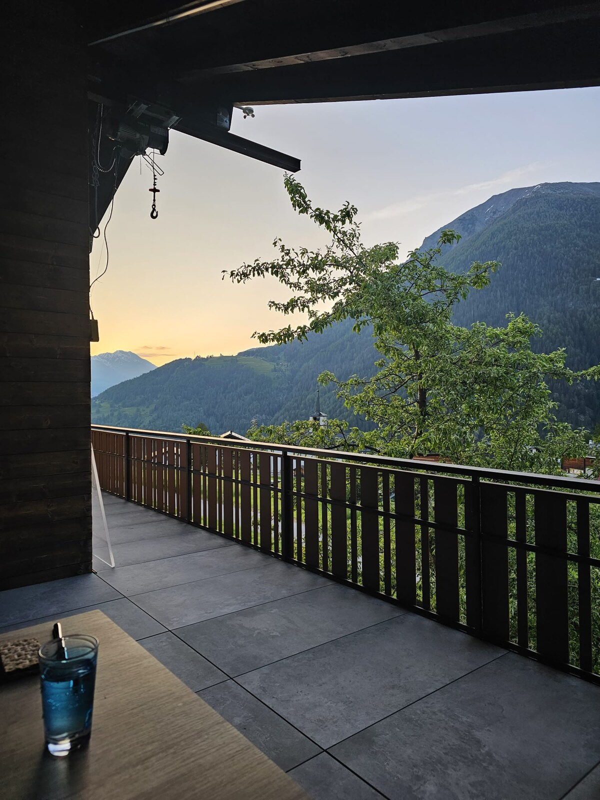 A spacious terrace is presented with a wooden railing, overlooking expansive mountain views bathed in soft sunset light. A table with a glass of water sits in the foreground, inviting relaxation amidst the natural surroundings.