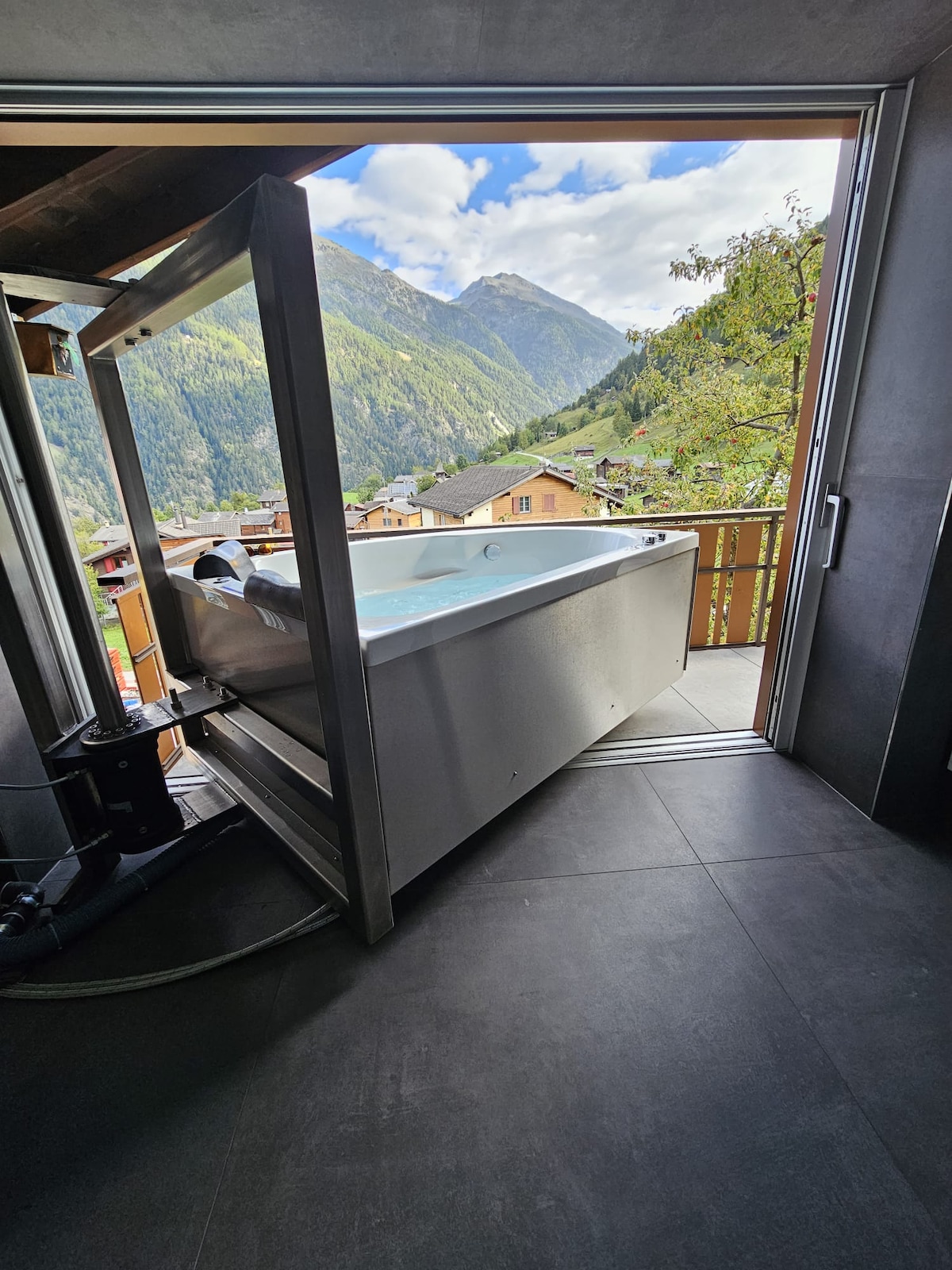 A sleek, modern bathtub extends from an open door, offering a direct view of the surrounding mountains. Natural light floods the space, highlighting the clean lines and contemporary design. The seamless transition between indoor and outdoor elements invites relaxation amidst scenic beauty.