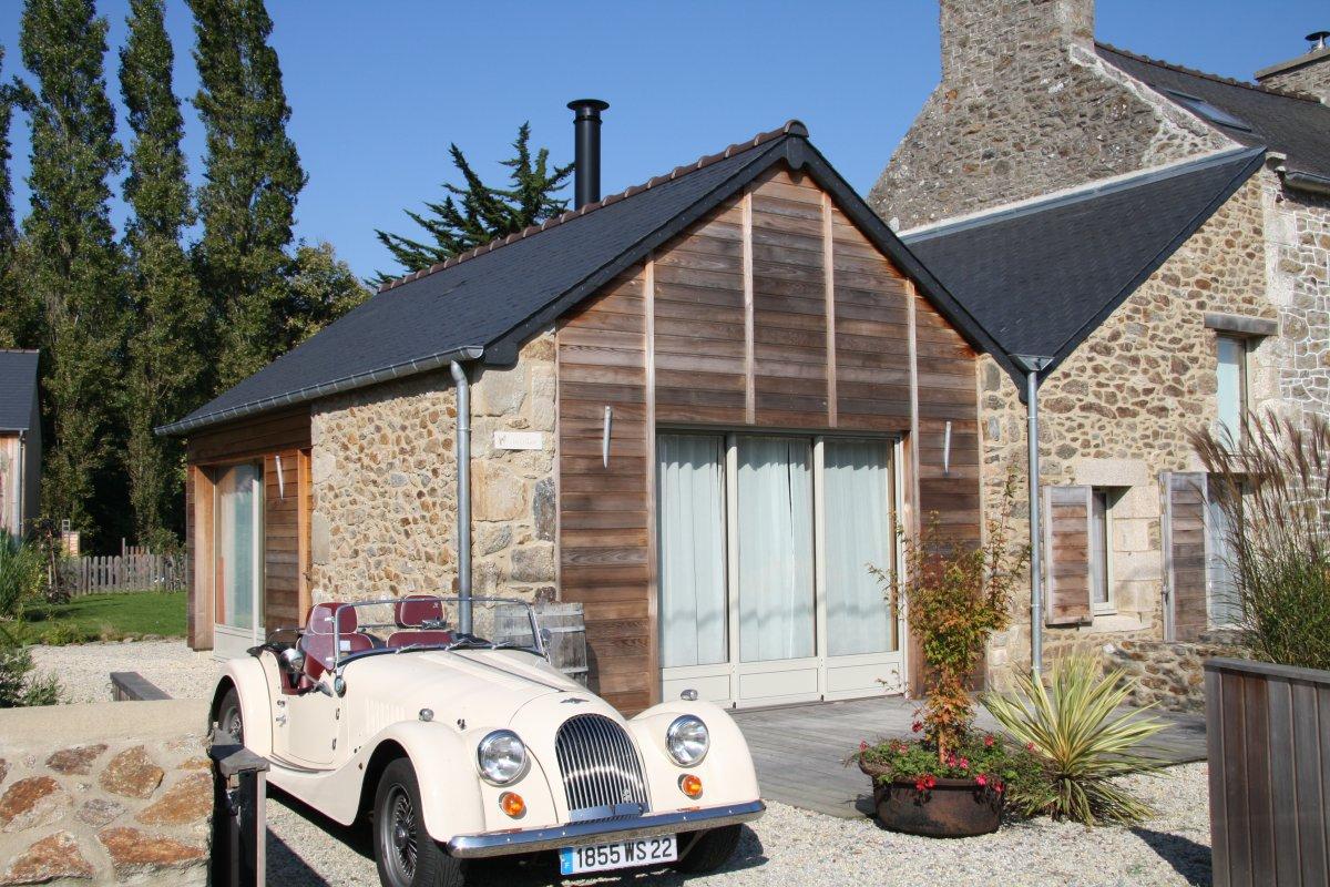 A charming stone and wood gîte is depicted, featuring large glass doors that open to a wooden terrace. A vintage car is parked in the foreground, with landscaped gravel and greenery enhancing the welcoming entrance.