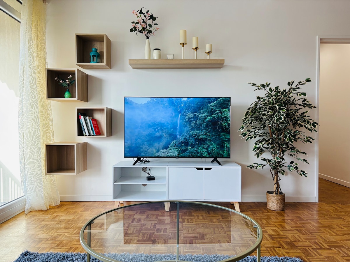 A bright living area is presented with a modern white TV stand beneath a large Smart TV displaying a scenic nature scene. A round glass coffee table sits on a plush rug, while decorative shelves showcase plants and candles, contributing to a cozy atmosphere.