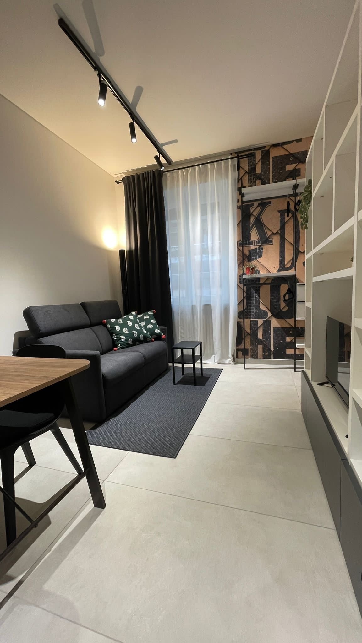 A modern living area features a sleek black sofa adorned with decorative pillows, positioned against a patterned wall. A small black table complements the sofa, while a light streams through sheer curtains, casting soft shadows on the tiled floor.