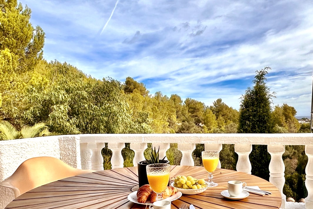 A wooden table is set for breakfast on a terrace, featuring a glass of orange juice, a small cup, a croissant, and a plate with fruit. The surrounding greenery is visible, with a scenic sky overhead, complemented by white balustrades.