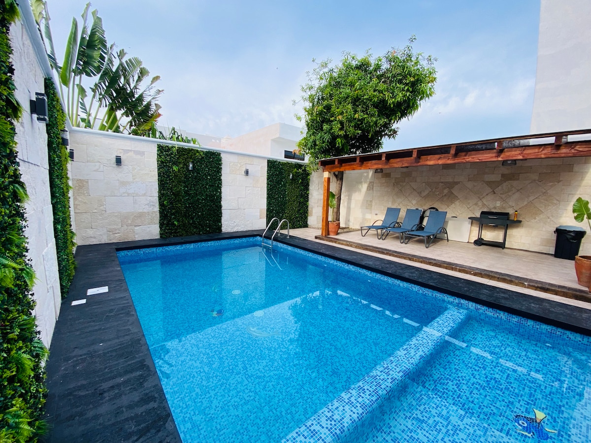 A private, heated pool is framed by lush greenery and a stone wall. Lounge chairs are positioned nearby, and a shaded patio area with a barbecue grill is visible. The tranquil water's surface reflects the sky above, inviting relaxation.