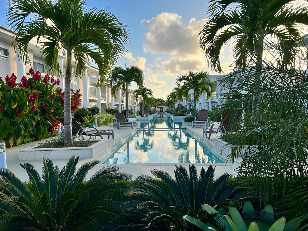 A tranquil pool area is surrounded by lush palm trees and vibrant flowering plants. Lounge chairs are positioned along the water's edge, creating an inviting space for relaxation. The pool reflects the sky and greenery, enhancing the serene atmosphere of the complex.