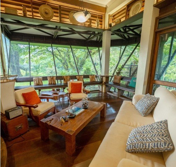 A spacious living area features large windows framing a natural view of the surrounding trees. Comfortable seating is arranged around a wooden coffee table, accented with decorative pillows and cushions. Light streams in, enhancing the relaxed atmosphere of the space.