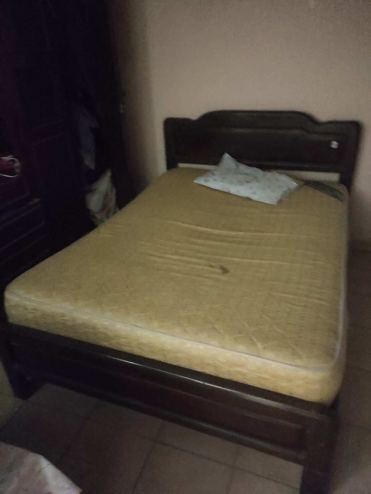 A wooden bed frame is shown with a lightly used mattress. A small decorative pillow is placed on the mattress. The surrounding walls have a neutral color, and a closet is visible in the background, providing additional storage space.