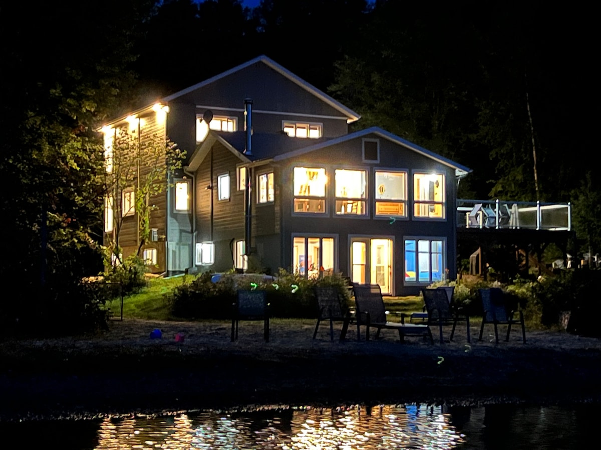The chalet is illuminated at night, showcasing its spacious design. Large windows create a warm glow, reflecting on the water's surface. Outdoor seating is visible in front, providing a comfortable gathering space overlooking the sandy beach and serene lake.